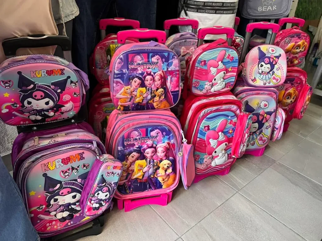 Mochilas Escolares Personalizadas 2027 2 se exponen coloridas mochilas escolares y bolsas para el almuerzo con personajes de dibujos animados, entre los que se incluyen unicornios y niñas de dibujos animados. La mayoría presenta tonos rosas y morados con diseños llamativos.